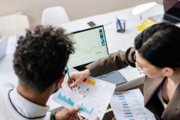 Two colleagues in a meeting room discussing financial charts and graphs on a laptop and paper.