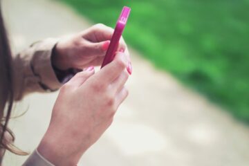 Woman holding a pink smartphone with both hands in a casual outdoor setting.