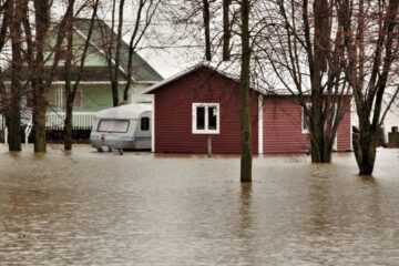 House and caravan surrounded by floodwaters among leafless trees in a natural setting.